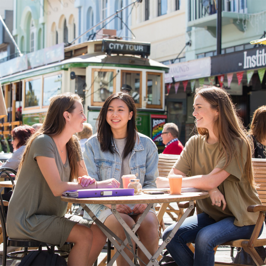 Three students sitting at a cafe table in New Regent Street with tram in the background